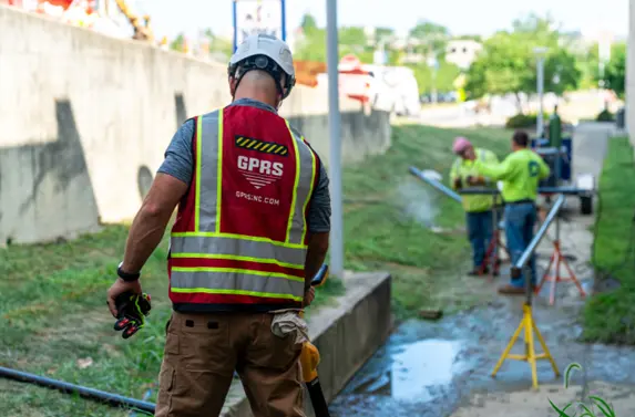 A GPRS Project Manager in the field, using an EM locator to find buried utilities on a jobsite. There is standing water in front of him beside a concrete pediment, a concrete wall to his left, and a sidewalk with two construction workers and equipment in the background. 