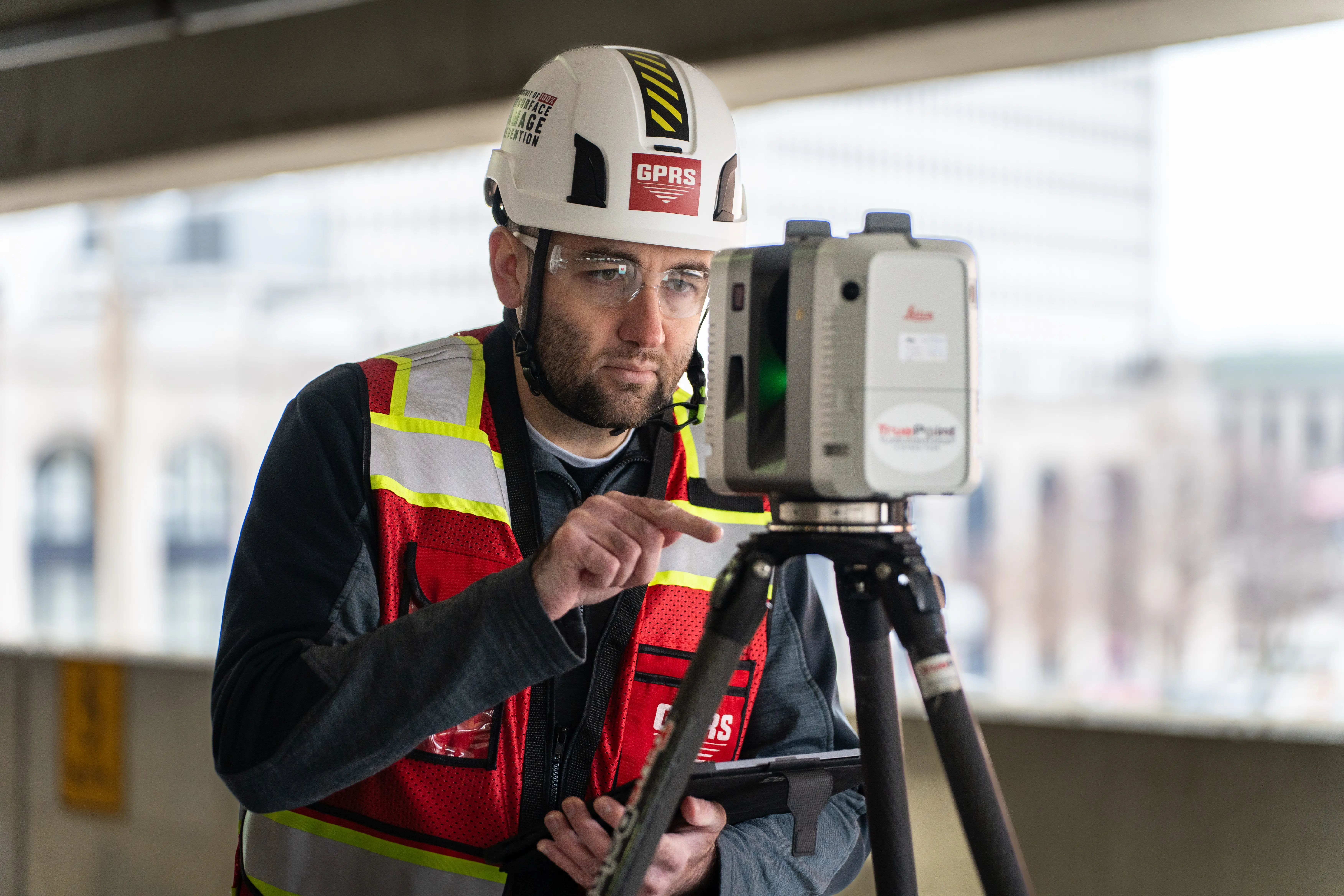 GPRS Project Manager wearing a white hard hat and red safety vest operates a 3D laser scanner mounted on a tripod inside a building.
