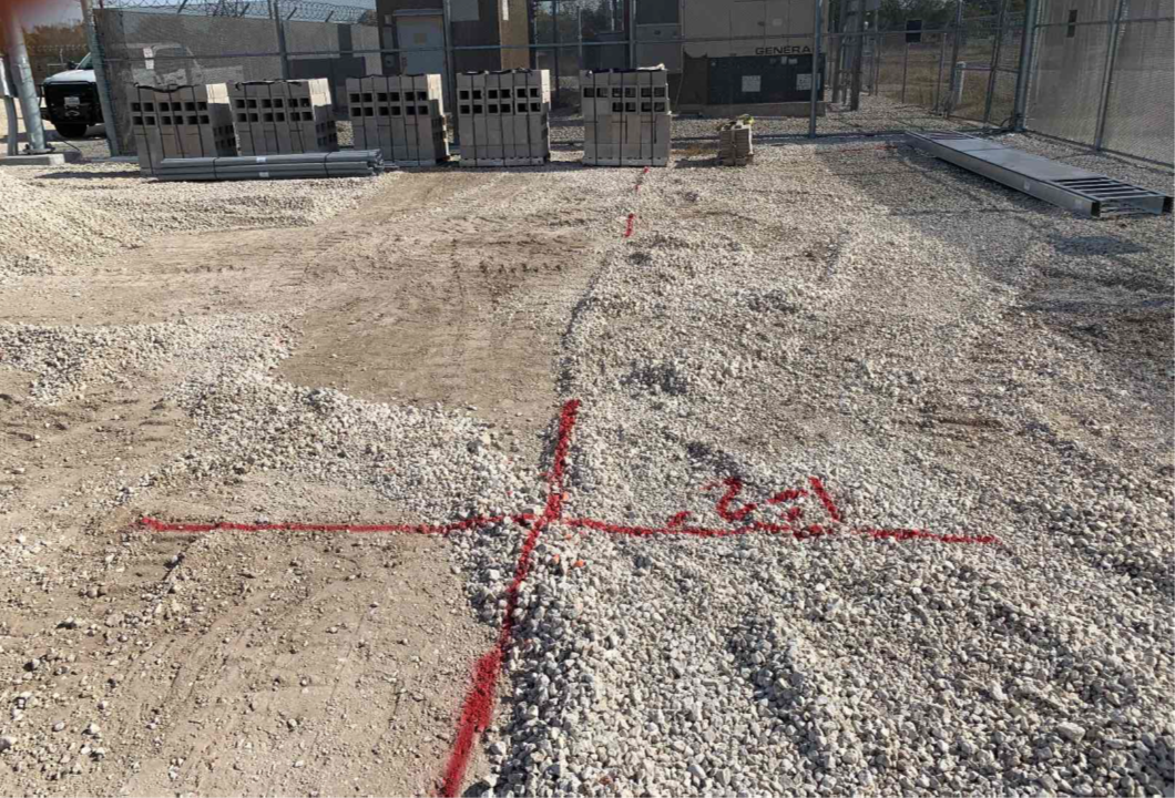 A dirt and gravel site with power transformers in the background. On the ground are some red lines in the shape of a cross made from red spray paint. Also written in red spray paint within the red cross is “1’-2’.”