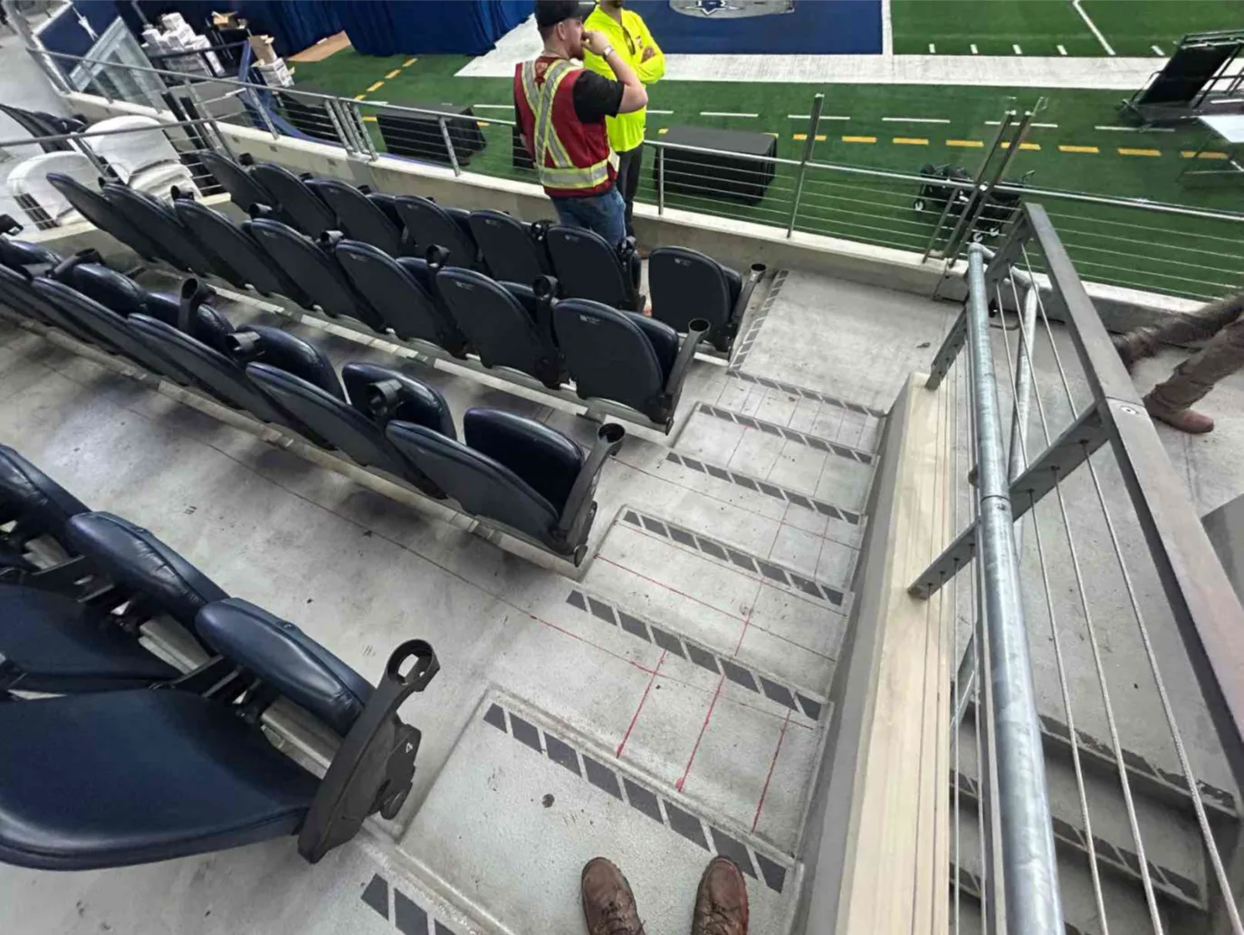 Seating areas with concrete floors and stairs that have red lines marked out to represent subsurface rebar. A GPRS Project Manager stands at the bottom row of the seats wearing a high visibility vest, jeans, and a black baseball hat. Beyond the seats is a portion of the Dallas Cowboys field and endzone. 