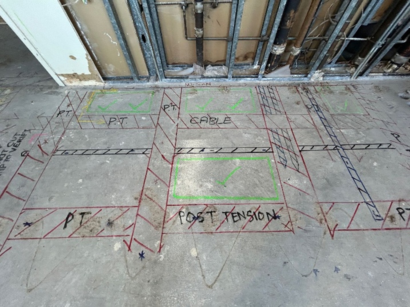 A concrete floor with visible piping on the wall in the background. On the slab are red, black, and green markings. The red lines consist of perpendicular lines with multiple diagonal lines in between that represent post tension cables. There are similar markings in black but of smaller size. There are also words written in black like, “PT CABLE,” and “POST TENSION.” There are four green boxes with green check marks representing GPRS’ Green Box Guarantee. 