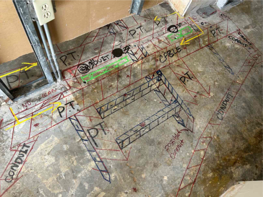 A concrete floor with a wall under construction in the background in the background. On the slab are red, black, and green markings. The red lines consist of perpendicular lines with multiple diagonal lines in between that represent post tension cables. There are similar markings in black but of smaller sizes. There are also words written in black like, “PT CABLE,” and “POST TENSION.” There are four green boxes with green check marks representing GPRS’ Green Box Guarantee.