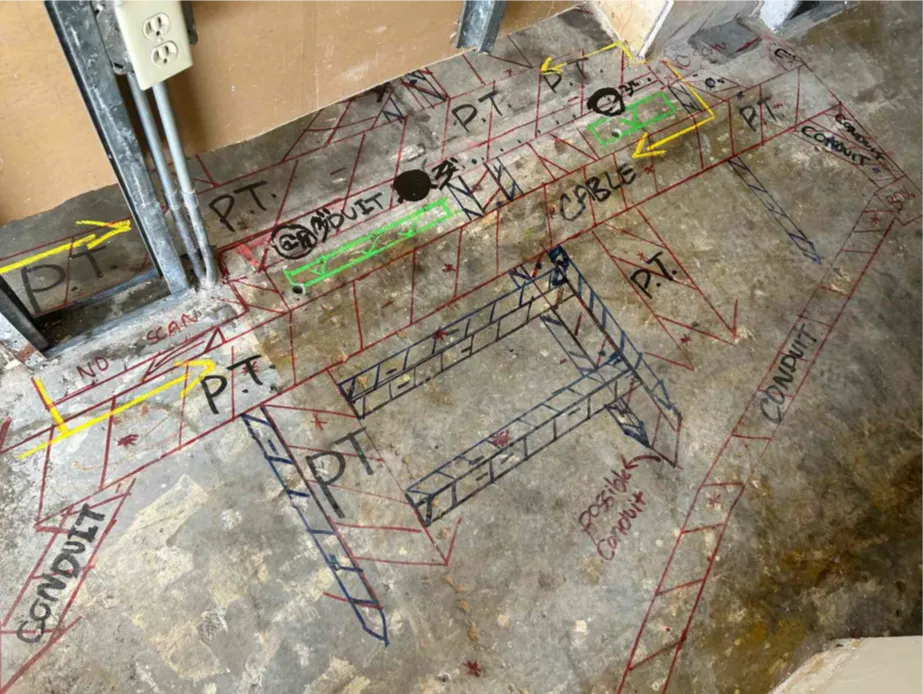 A concrete floor with a wall under construction in the background in the background. On the slab are red, black, and green markings. The red lines consist of perpendicular lines with multiple diagonal lines in between that represent post tension cables. There are similar markings in black but of smaller sizes. There are also words written in black like, “PT CABLE,” and “POST TENSION.” There are four green boxes with green check marks representing GPRS’ Green Box Guarantee.