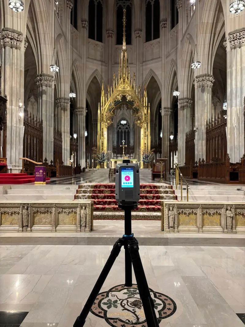 GPRS Project Manager wearing a high‑visibility vest and operating a tripod-mounted 3D laser scanner on the front steps of the St. Patrick’s Cathedral with tall towers, ornate architectural details, and a large circular stained‑glass window.