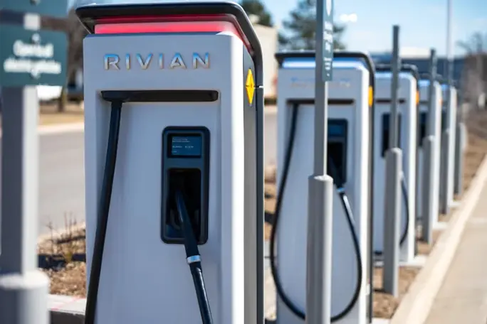 A row of Rivian electric vehicle charging stations is shown along a curbside area, with charging cables attached and additional stations visible in the background.