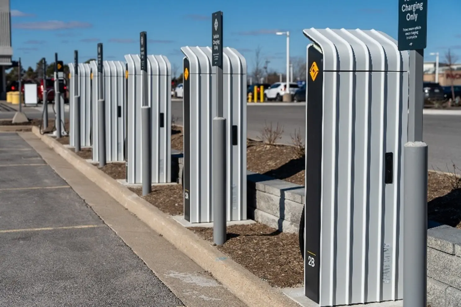 A row of modern electric vehicle charging stations lines a parking area, each with a tall, ribbed exterior design and accompanying signage, set against an open lot with buildings and trees in the background.