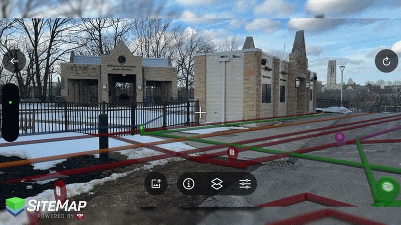 Augmented reality overlay showing underground utility lines in multiple colors (red, orange, purple, and green) aligned across a paved roadway near a gated stone entrance, with snow on the ground and leafless trees in the background.