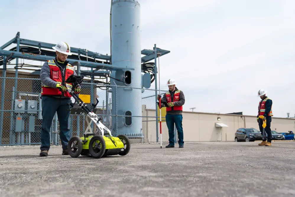 Three GPRS Project Managers wearing red high visibility vests with hard hats performing utility locating services at a telecom tower site. One is on the left side pushing a neon yellow GPR pushcart. One in the middle is collecting RTK-positioned satellite data. The one on the far right is collecting data using an EM locator.