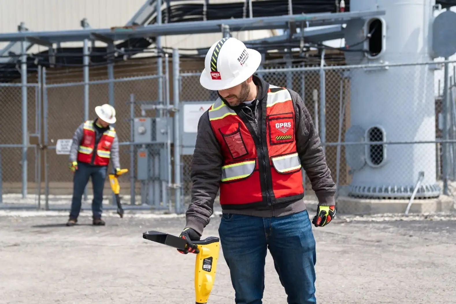 Two GPRS Project Managers collecting utility data with EM locators while wearing red high-visibility vests and hard hats on a telecommunications tower site.