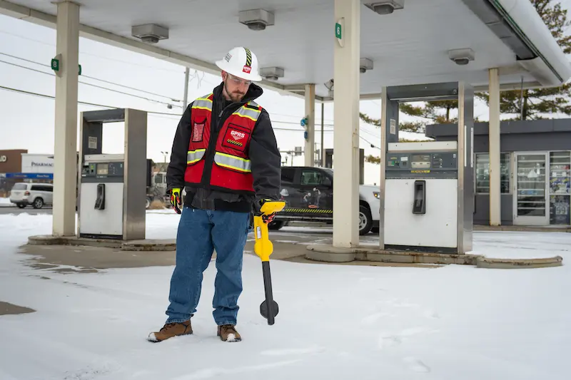 GPRS Project Manager in a high-visibility safety vest and hard hat stands in snow under a gas station canopy, holding an electromagnetic (EM) locator near fuel pumps and a parked car.