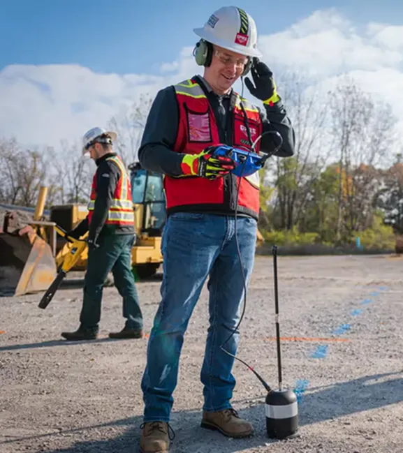 A GPRS Project Managers wearing safety gear, a hard hat, and a high visibility vest operates an acoustic leak detector with an elephant’s foot attached on a jobsite, while another Project Manager in similar protective gear scans the area using an EM locator.