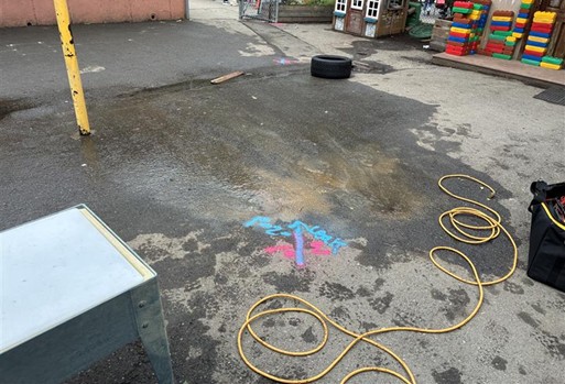Wet asphalt surface with red and blue spray paint markings indicating underground utilities, surrounded by a coiled yellow cable, a loose tire, a metal utility box, and nearby playground equipment in an outdoor area.