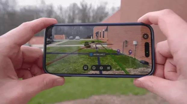 Red brick buildings with a grassy lawn in the foreground on an overcast day. Two Caucasian hands hold an iPhone to reveal green, blue, red, and purple lines over the grass, representing buried utilities in augmented reality. 