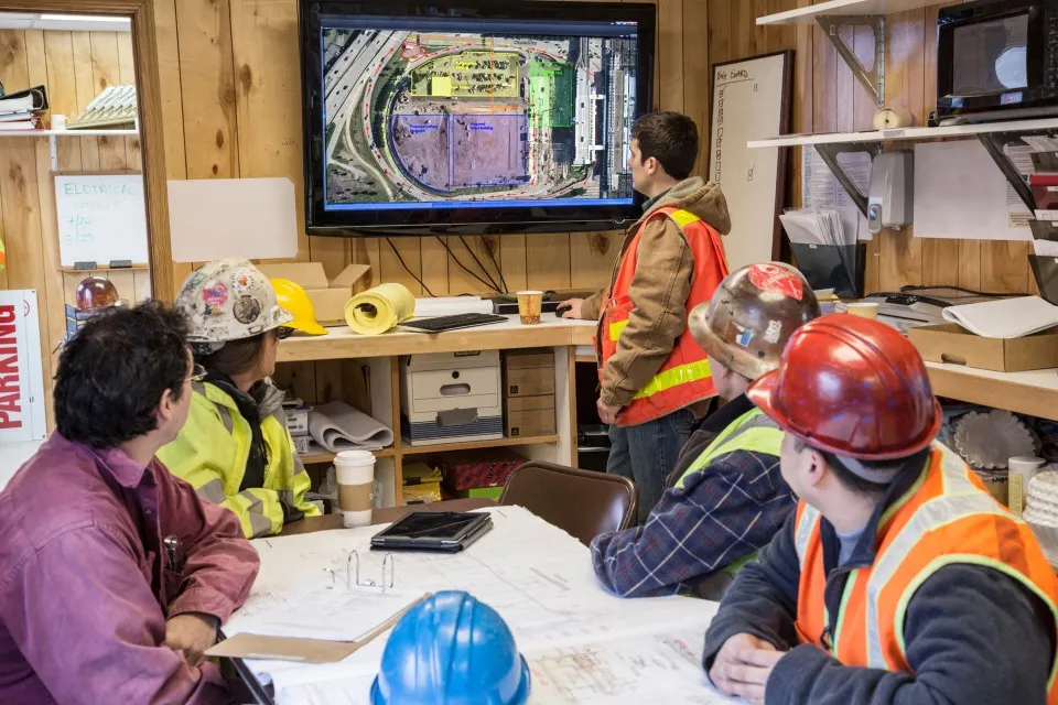 A group of construction workers wearing safety vests and hard hats sit around a table in a site office while one person stands and points to a large screen displaying an aerial construction map, as others look on with plans and documents spread across the table.