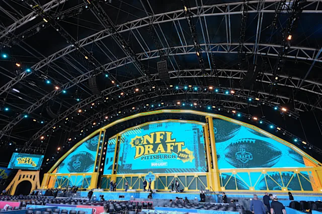 Wide view of a large outdoor stage set up for the NFL Draft in Pittsburgh, featuring a massive curved LED screen with “NFL Draft Pittsburgh 2026” displayed in blue and yellow. Metal trussing and stage lighting arch overhead, with rows of seating and people visible in front of the stage.