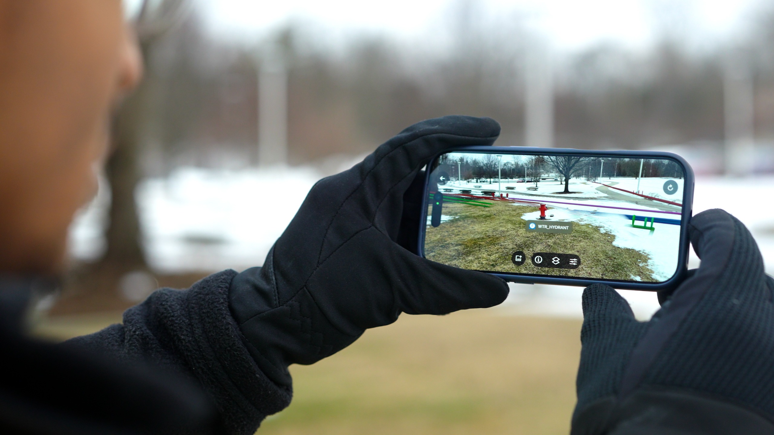 A person wearing gloves and holding a smart phone that shows the POV of the phone's camera with the underground utilities populating above the ground and next to the red fire hydrant.