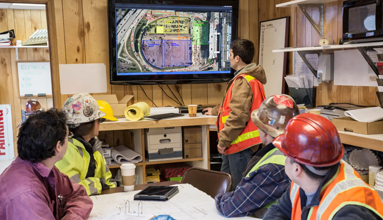 Four men in construction gear stare at the whiteboard to discuss the daily operations on site