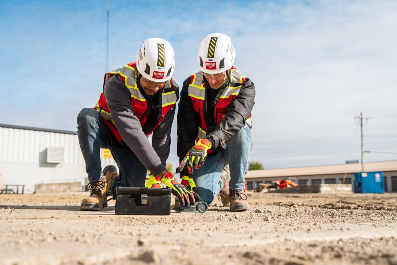 Two men in high-viz gear use ground penetrating radar to scan concrete.