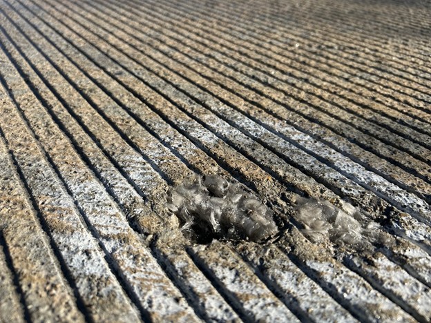 A close-up shot of a sample embedded in a bridge surface shows a visible clump of synthetic reinforcement fibers, a fiber ball defect, embedded within the concrete. The bridge deck and surrounding road are visible in the background.