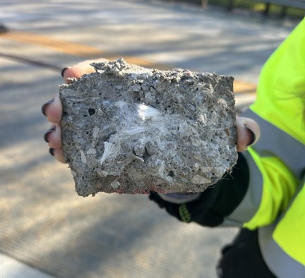 A close-up of a bridge topping slab surface showing a dense clump of synthetic concrete reinforcement fibers protruding through the grooved concrete deck. The fiber ball defect appears as a rough, tangled mass pushing up through the surface.