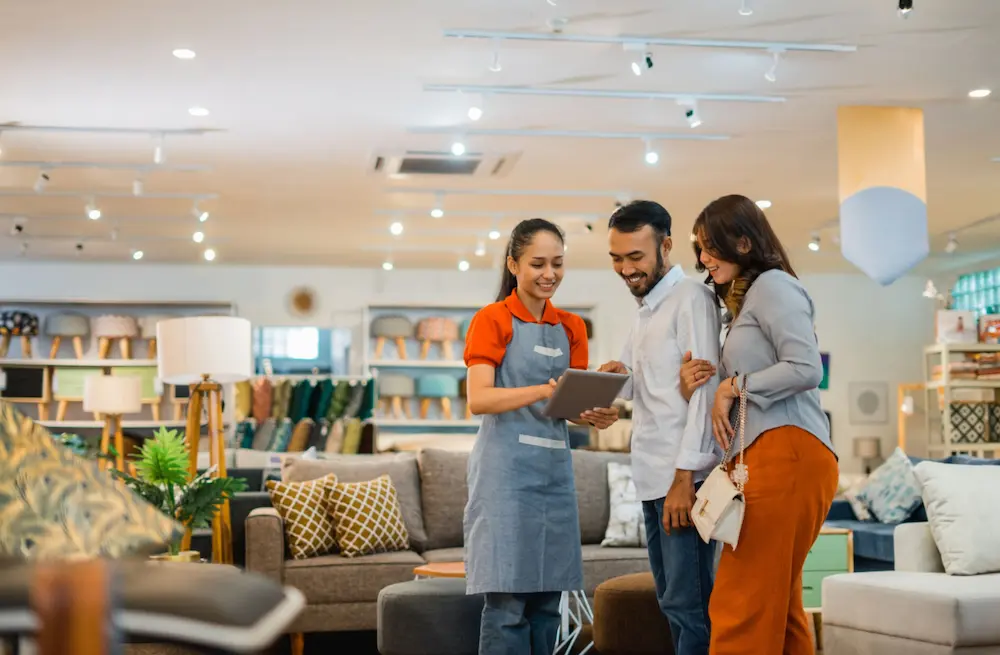 A female clerk with an iPad helps a couple inside a furniture store.