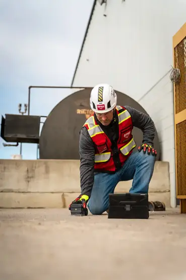 A GPRS Project Manager is down on one knee while scanning a concrete slab outside a building. A storage tank and a large yellow metal safety door are behind him. He is wearing proper PPE: vest, hard hat and cloves as he scans. 