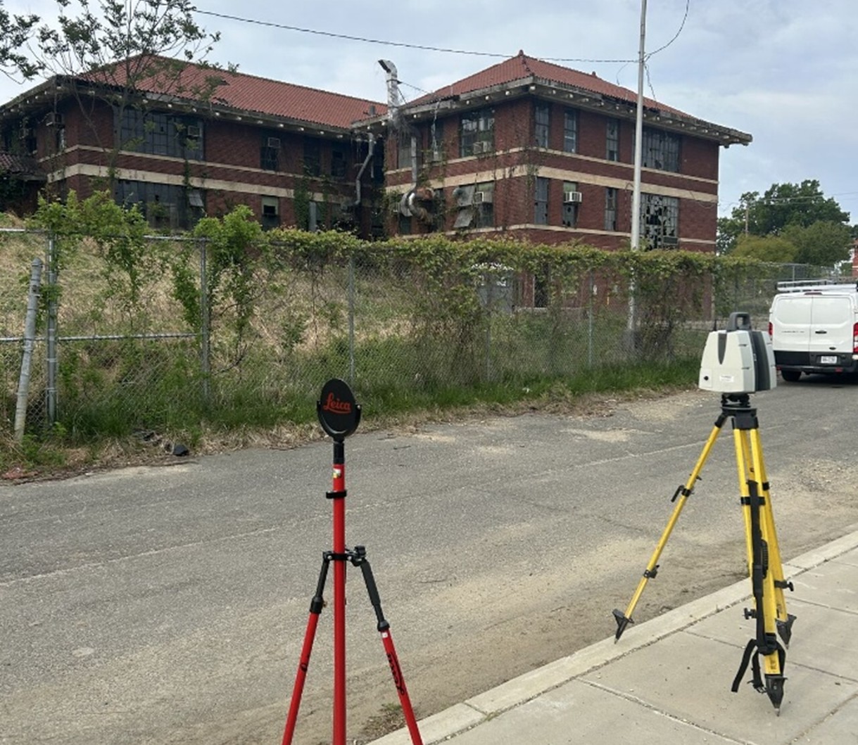 Street-level view of an older two-story red brick building with a red tile roof and rows of tall windows, many of which appear broken or missing. A chain-link fence covered with overgrown vines runs along the front of the property. In the foreground on the sidewalk and street, two surveying instruments are set up on tripods, one red Leica target and one gray scanning device on a yellow tripod. A white van is parked on the right side of the frame. The scene appears overcast, with muted lighting and a slightly rundown environment.