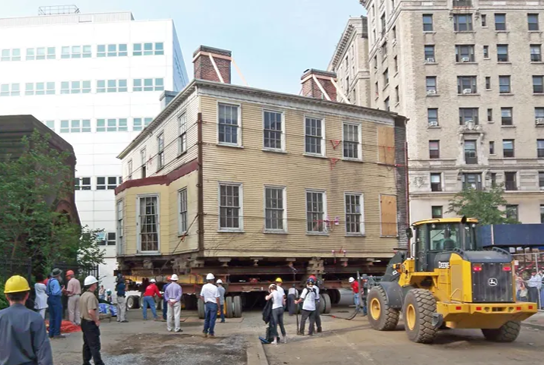 A historic red-brick building with boarded-up arched windows is elevated on a specialized moving platform at a construction site. Large yellow cranes and lifting equipment surround the structure, while a multi-story building under construction rises in the background beneath a partly cloudy sky.