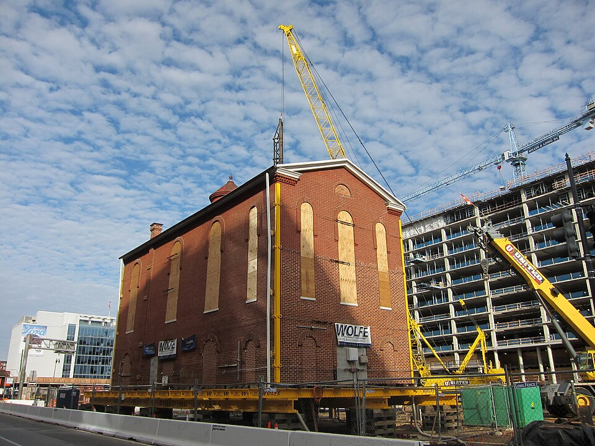 A historic red-brick building with boarded-up arched windows is elevated on a specialized moving platform at a construction site. Large yellow cranes and lifting equipment surround the structure, while a multi-story building under construction rises in the background beneath a partly cloudy sky.