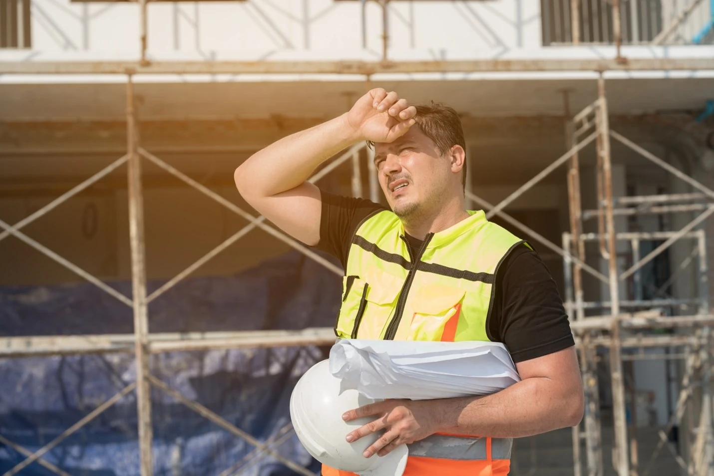 Construction worker overheated on a job site during summer heat, holding a white hard hat and rolled blueprints in front of scaffolding.
