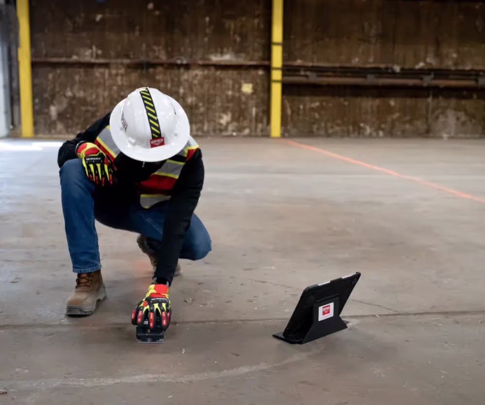 Worker wearing safety gear scans concrete floor with handheld device while a tablet displays data nearby.