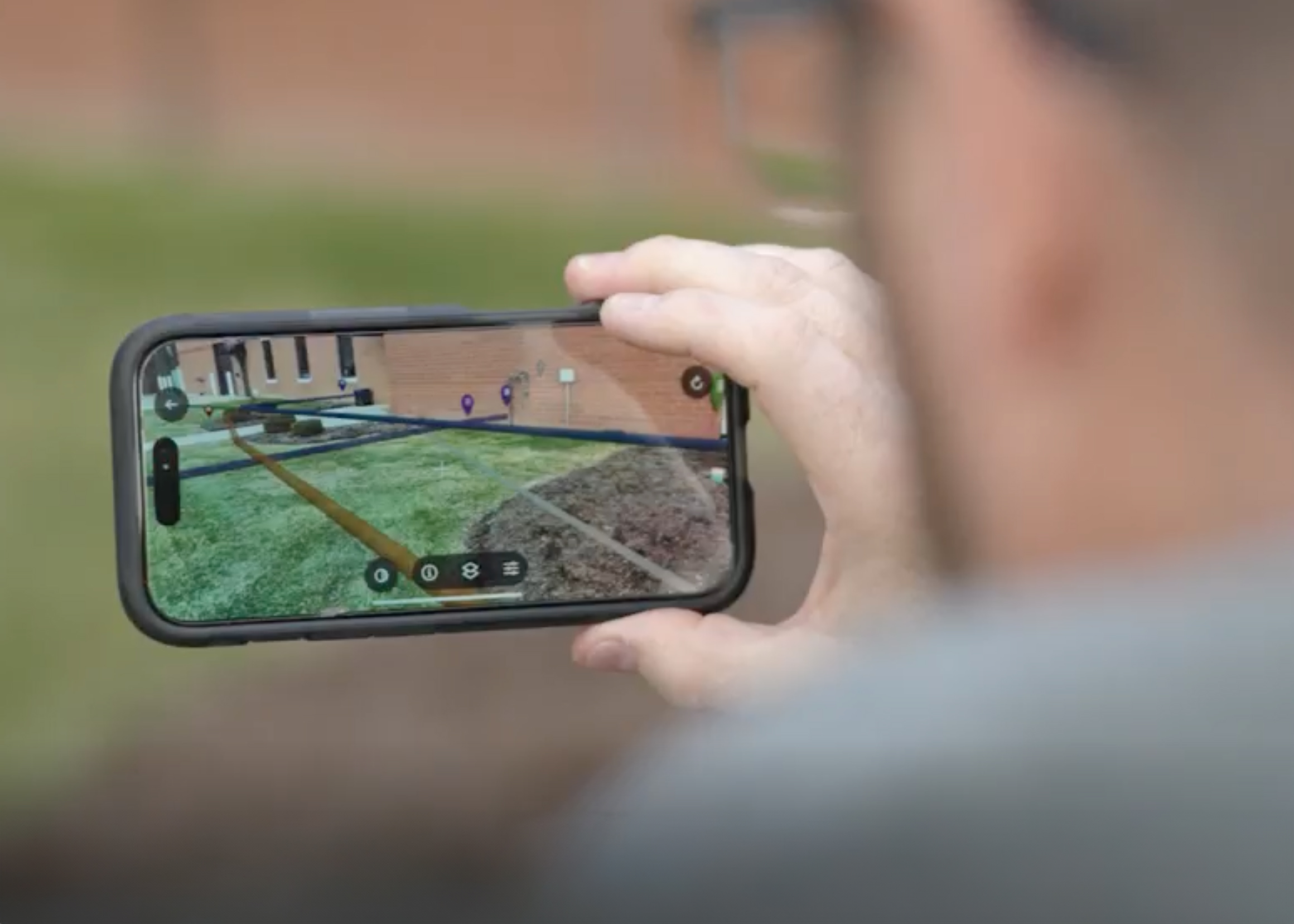 A person holds up a smartphone outdoors, showing an augmented reality overlay of underground utilities and pipe paths aligned with the real-world environment.