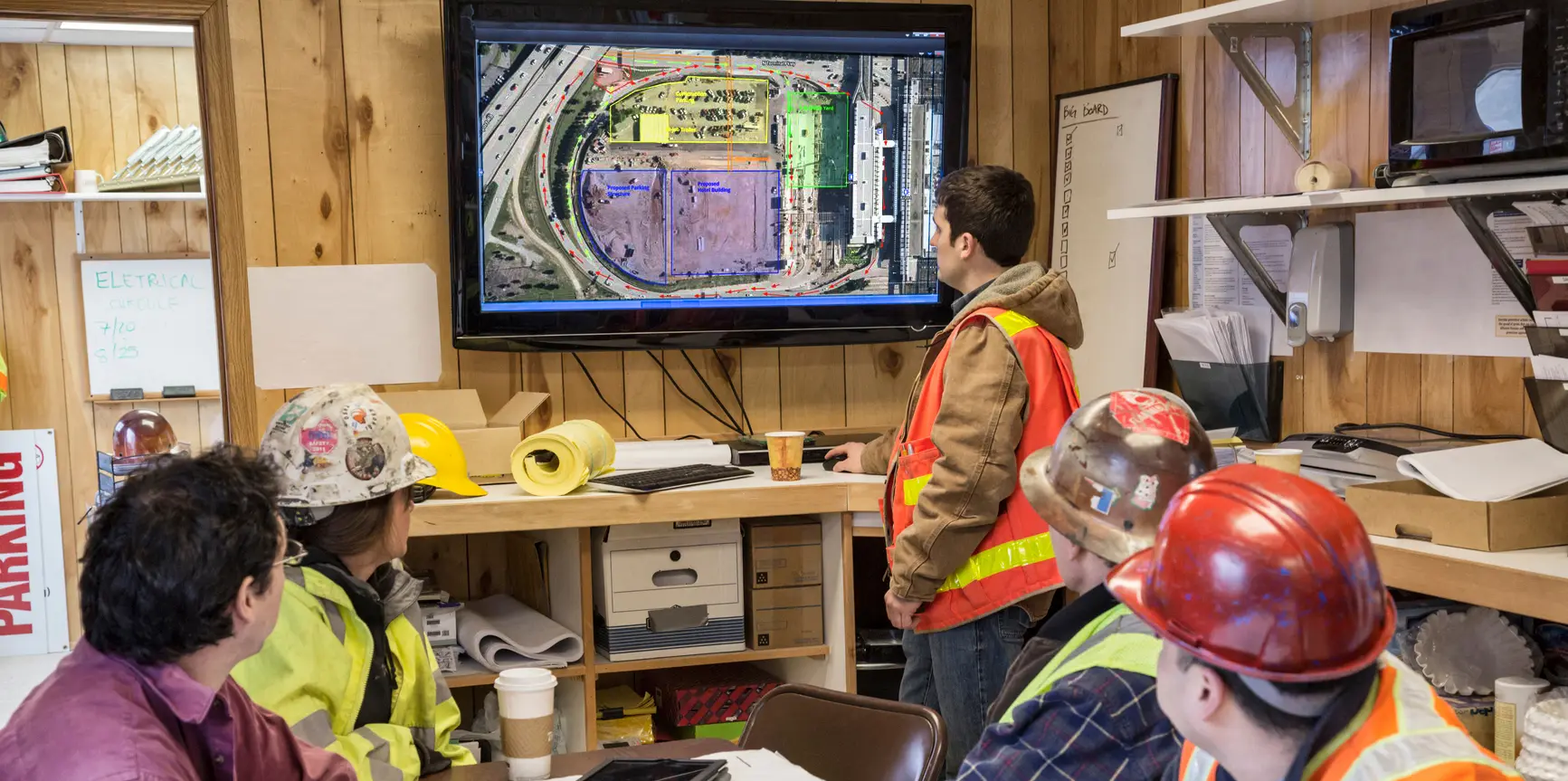 Construction crew inside a jobsite trailer watches a display screen showing a digital excavation map. Workers wearing safety vests and helmets are seated while one person stands near the screen.