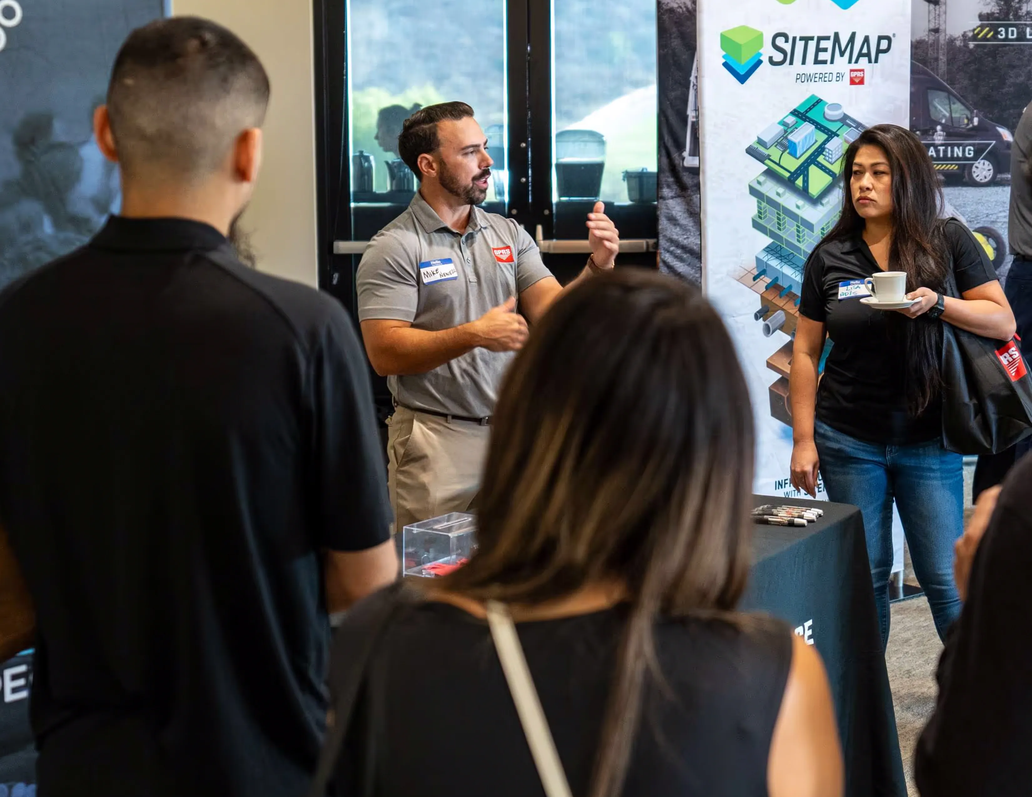 A man in a grey shirt and blue lanyard is talking to a woman in a black shirt.