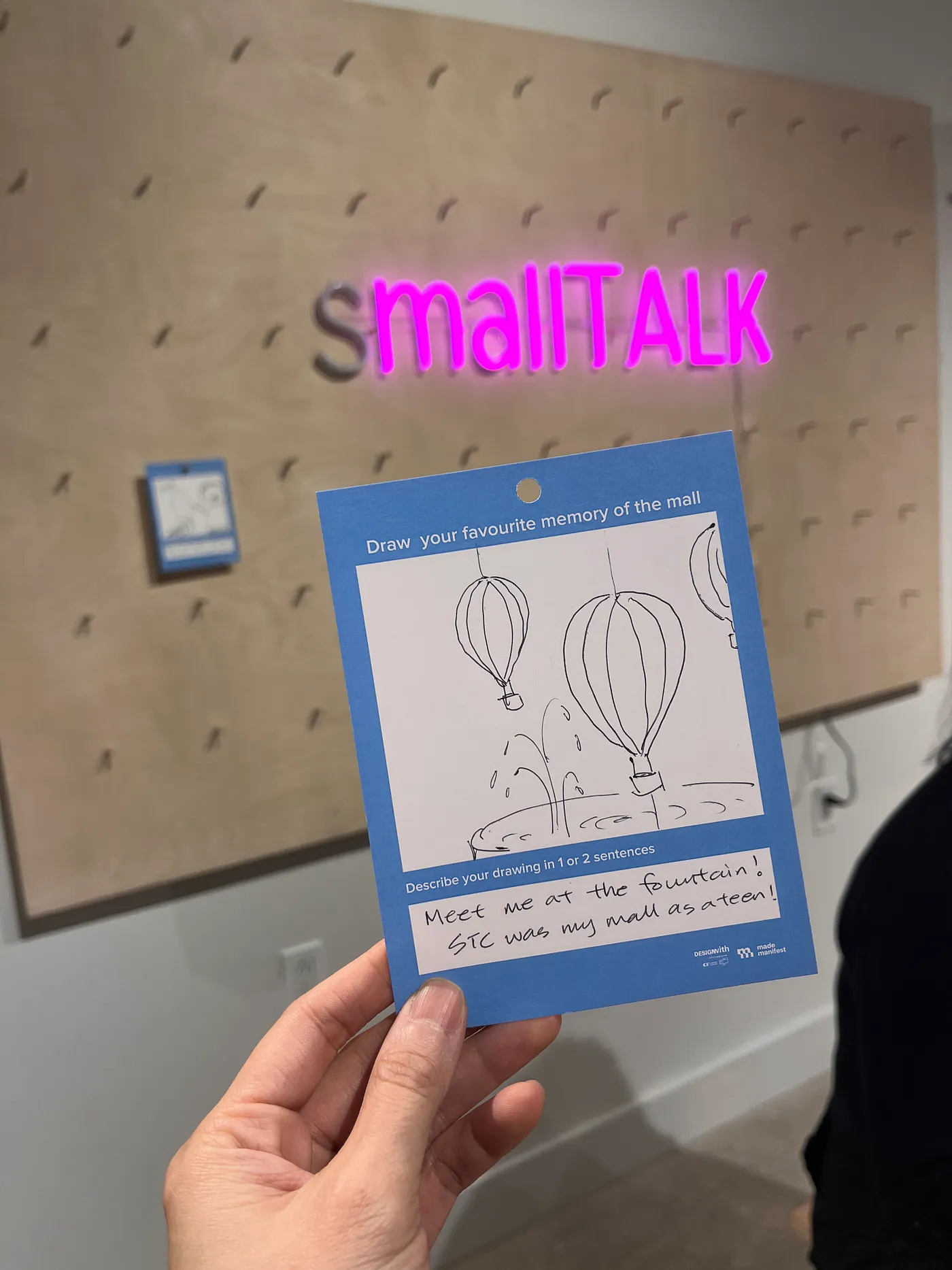 a blue card with the prompt draw your favourite memory of the mall. Someone has drawn the Scarborough Town Centre mall fountain which has hot air balloons in it. In the background, a neon sign on a pegboard reads 'smallTALK'.