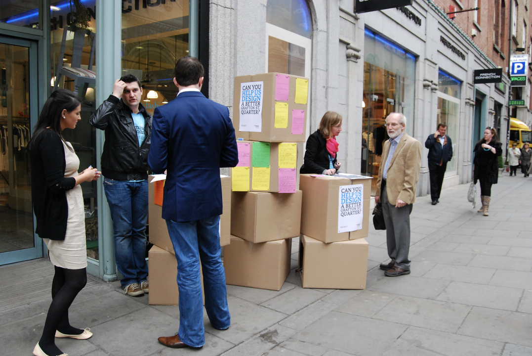Two researchers standing behind a stack of cardboard boxed with posters reading 'help us design a better grafton quarter' in Dublin. Each researcher is talking to a member of the public.