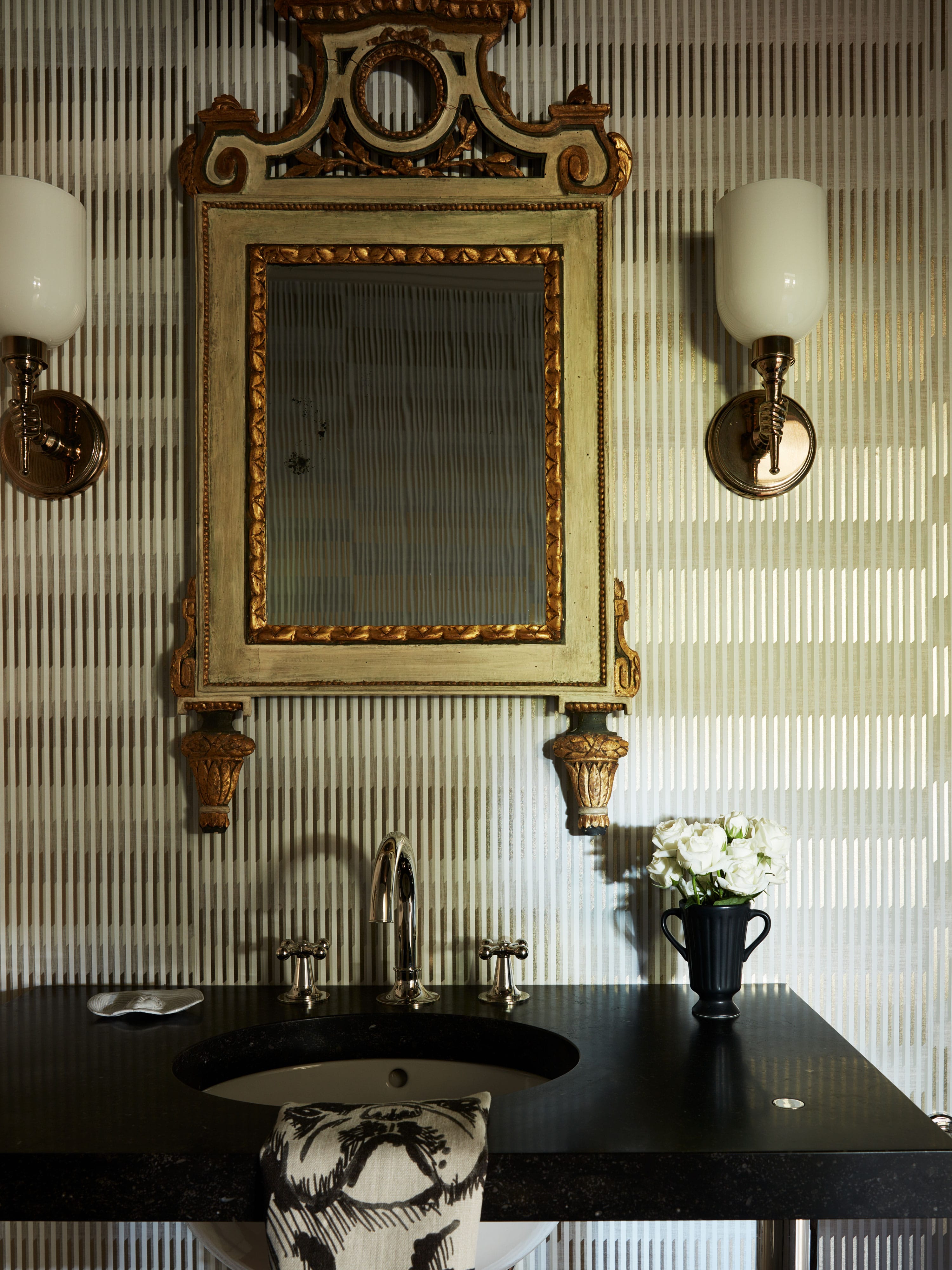 Bathroom sink with black countertop, ornate gold-framed mirror, wall sconces, and black vase with white flowers.