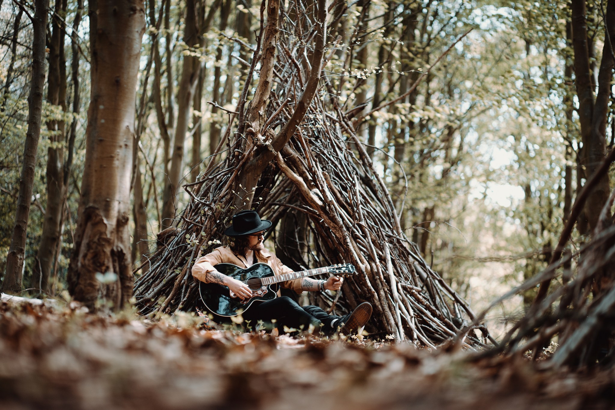 A musician sits in a forest, a wigwam of sticks behind him