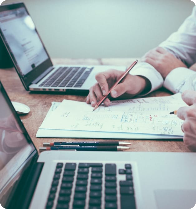 A person at their desk writing on a pad of paper with a Macbook