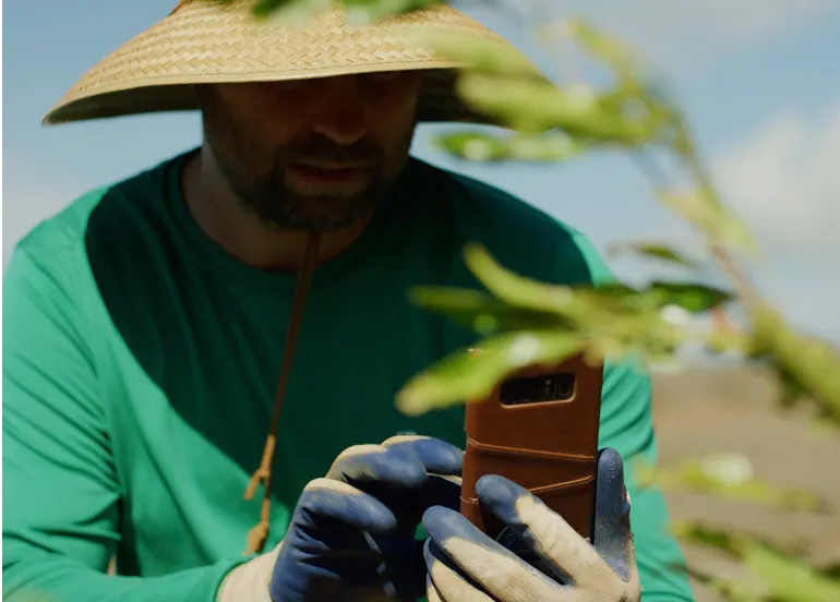 Person in a green shirt and wide straw hat using a smartphone outdoors near plants.