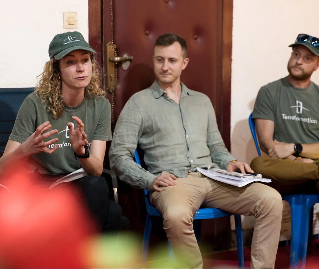 A woman wearing a green Terraformation cap and t-shirt speaks animatedly, gesturing with her hands, while two men listen attentively, seated on blue chairs indoors.