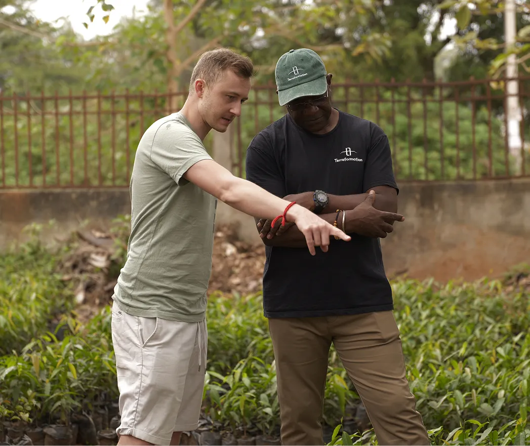 Two men standing outdoors in a garden or nursery with rows of small plants; a man in a light green t-shirt and beige shorts points towards something while talking to a man wearing a green cap and black t-shirt with the logo and text 'Teraformation'.