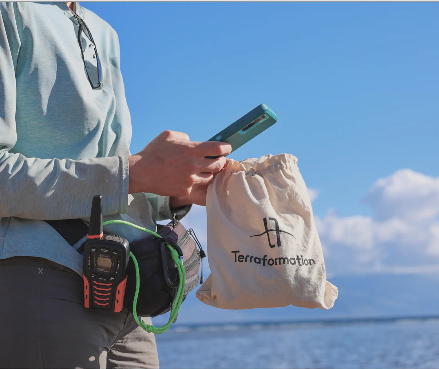 Person wearing light blue long sleeve shirt and gray pants stands near water holding a beige drawstring bag with the Terraform logo in one hand and a green smartphone in the other, with a walkie-talkie and pouch attached to their waist, under a clear blue sky with some clouds.