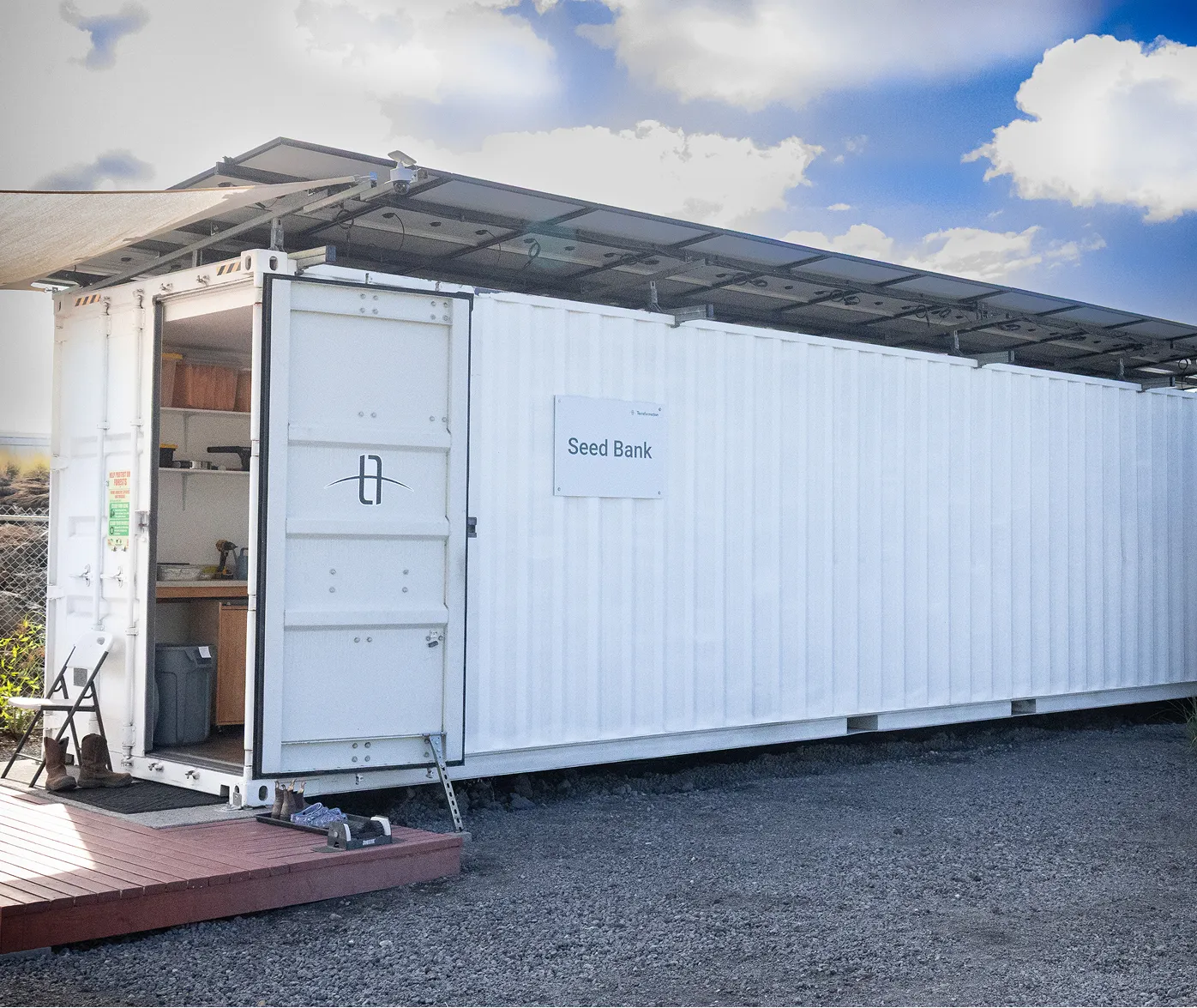 A large white shipping container labeled 'Seed Bank' with an open door revealing interior shelves and equipment, solar panels installed on top, and boots and shoes placed outside on wooden decking and ground.