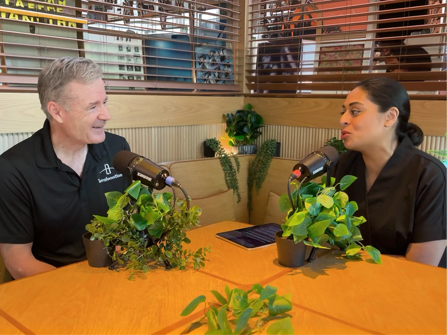Two people sitting at a wooden table in a booth with plants, speaking into Shure microphones during a podcast or interview.
