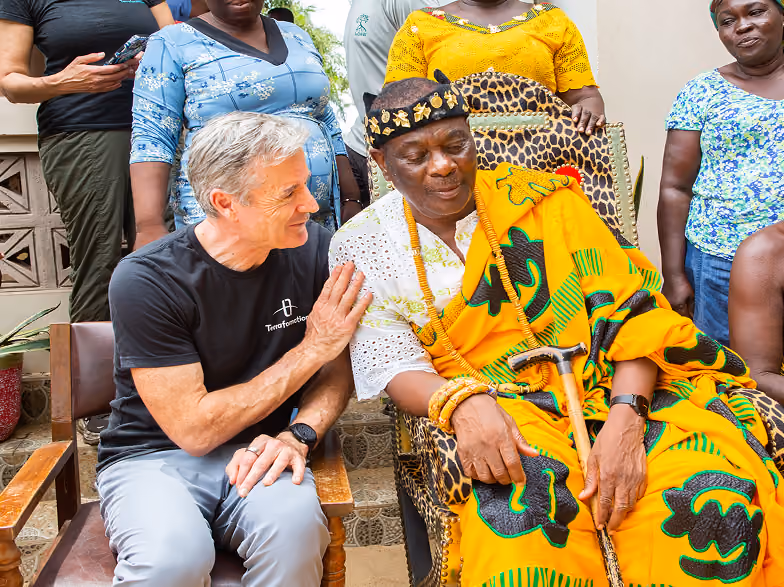 A man in a black Terraformation t-shirt sits next to an elderly man dressed in vibrant traditional African clothing and jewelry, holding a carved wooden cane, surrounded by several people standing in the background.