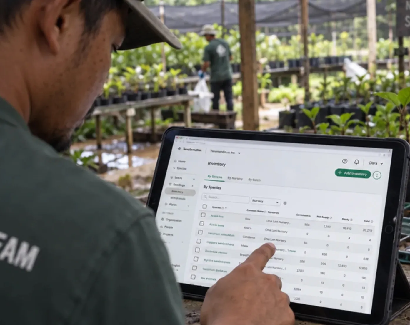 A person wearing a green shirt with partially visible text 'TEAM' points at an iPad screen displaying an inventory list of plant species in a nursery, with green plants and another person in the background inside a shaded plant nursery.