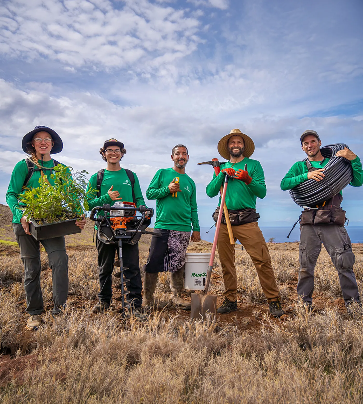 Five people in green shirts standing in a dry field under a partly cloudy sky, holding gardening tools and plants, smiling at the camera.