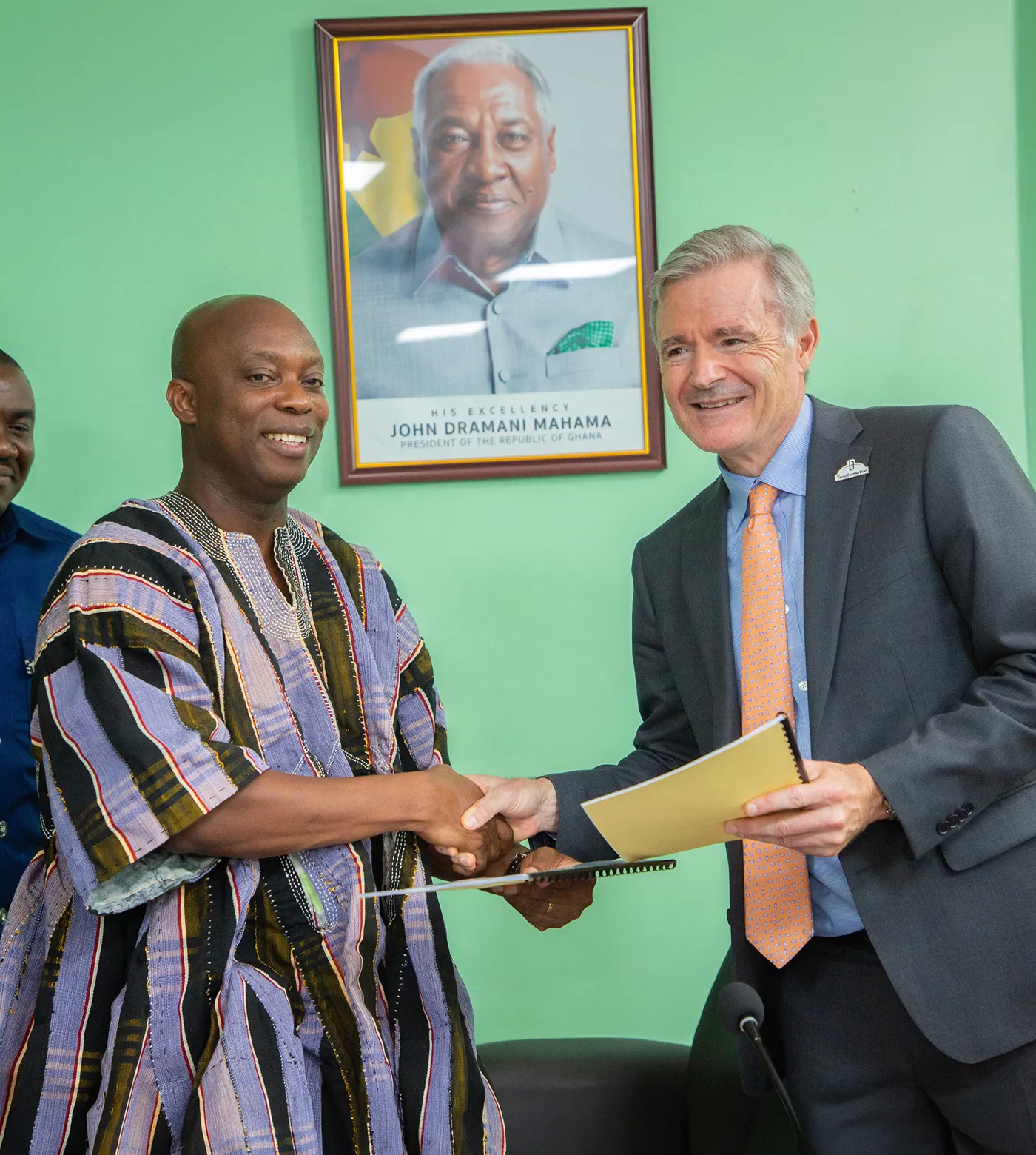 Two men smiling and shaking hands while holding documents in an office with a green wall and a framed portrait of John Dramani Mahama, former President of Ghana, in the background.