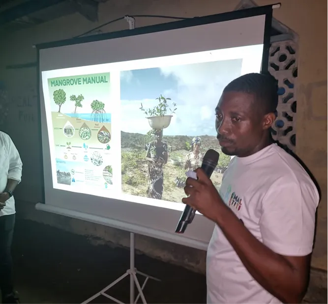 Man holding a microphone giving a presentation next to a projection screen displaying a 'Mangrove Manual' infographic and a photo of women carrying plants in baskets on their heads outdoors.